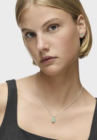 Turkusowy wisiorek na srebrnym łańcuszku, noszony z dopasowanymi kolczykami wkrętami; modelka ma krótkie, proste blond włosy i szary top.