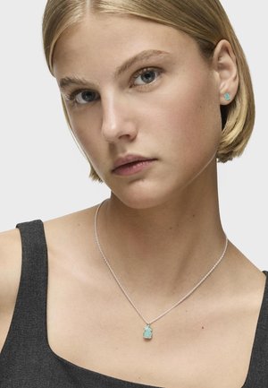 Turkusowy wisiorek na srebrnym łańcuszku, noszony z dopasowanymi kolczykami wkrętami; modelka ma krótkie, proste blond włosy i szary top.