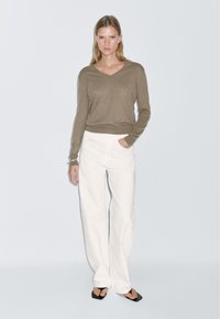 Brauner Strickpullover mit V-Ausschnitt, kombiniert mit weißen, weit geschnittenen Hosen. Verfügt über einen lässigen Schnitt und eine glatte Textur. Schwarze Sandalen runden das Erscheinungsbild ab.