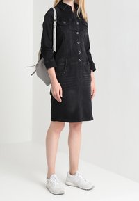 Svart denimklänning med knappfront, två bröstfickor och sidfickor. Tillsammans med vita sneakers och en ljusgrå ryggsäck.