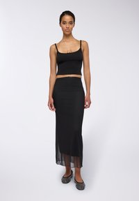 Top corto in maglia nera con spalline sottili, abbinato a una lunga gonna nera con uno spacco laterale e una finitura testurizzata. Slip-on neri scintillanti.