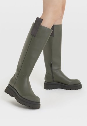 Knæhøje olivengrønne støvler med tykke sorte såler, sidelommer med lynlås og trækstropper, båret på bare ben mod en hvid baggrund.