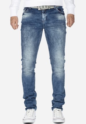 Mann trägt ausgewaschene blaue Slim-Fit-Jeans, weißen Gürtel, weiße Turnschuhe und ein weißes Langarmhemd, steht vor einem einfarbigen Hintergrund.