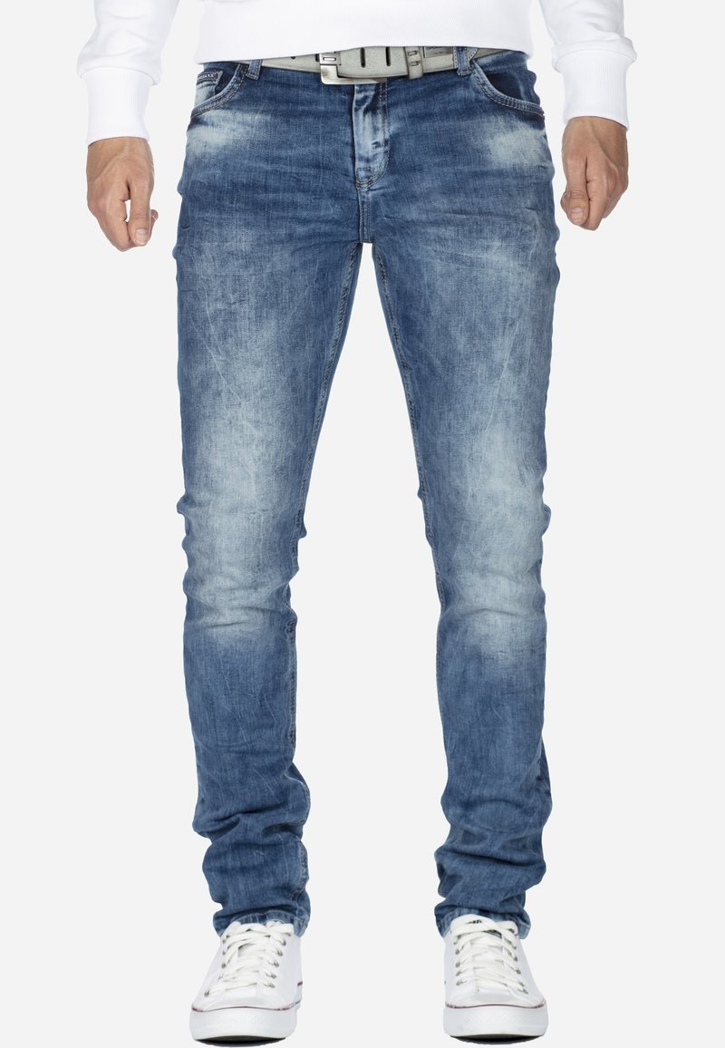 Homme portant un jean slim bleu délavé, une ceinture blanche, des baskets blanches et une chemise blanche à manches longues, debout devant un fond uni.