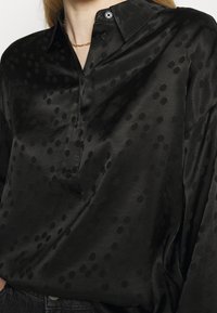 Svart satinblus med ett subtilt prickmönster, långa ärmar, krage och knappslå. Slät yta med en avslappnad passform.