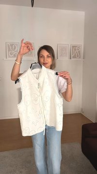 Fehér, ujjatlan felső hímzett virágmintákkal, magas nyakkivágással és elöl megkötő pántokkal. Egy személy által egy akasztón bemutatva.
