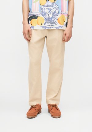 Μπεζ παντελόνι ίσιας γραμμής συνδυασμένο με καφέ δερμάτινα παπούτσια με κορδόνια και πολύχρωμο πουκάμισο με σχέδιο μπλε βάζου και κίτρινου φρούτου.