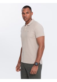 Beige polokošile, krátké rukávy, límeček a vyšitá nášivka na hrudi. Model stojí proti bílému pozadí, oblečený v tmavozelených kalhotách a hodinkách.