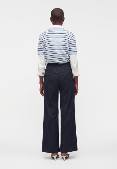 Personne debout de dos, portant un pull à manches courtes rayé bleu clair sur une chemise blanche à manches longues, un pantalon large à rayures bleu marine et des talons blancs.