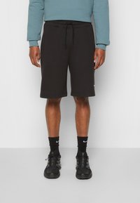 Černé šortky z měkké látky, s elastickým pasem na šňůrku a bočními kapsami. Nosené s černými ponožkami a teniskami.