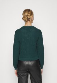 Žena s blond vlasy v drdolu, oblečená v tmavě zeleném texturovaném svetr a černých kožených kalhotách, zobrazená zezadu na jednoduchém pozadí.