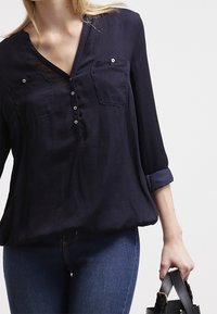 Mörkblå blus med v-ringning, två bröstfickor, knäppning med knappar, uppvikta ärmar och mjukt, flytande tyg. Bärs med åtsittande jeans och en väska.