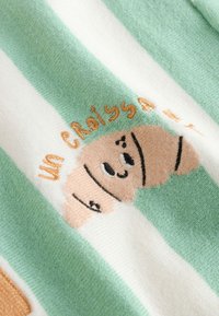 Tissu rayé vert et blanc avec un croissant souriant brodé et le texte « un Croissant » en fil orange.