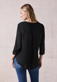 Sort bluse med lange ærmer, rund halsudskæring, afslappet pasform og plissering bagpå. Kombineret med blå jeans, vist fra bagenden.