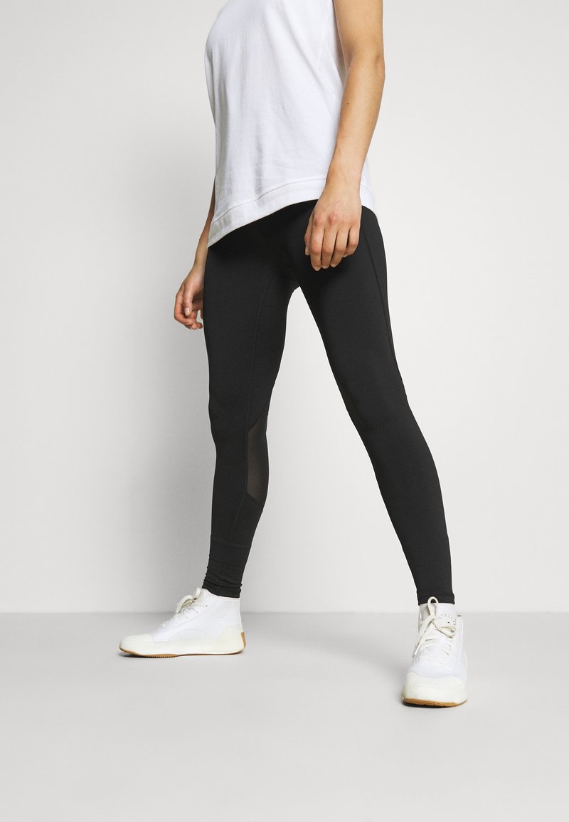 Svarta leggings gjorda av stretchigt material, med en åtsittande design, subtila meshpaneler, i kombination med en vit t-shirt och sportiga vita skor.