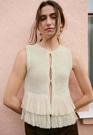 Joven con el cabello suelto, lleva un top de punto sin mangas color crema con volantes en capas, de pie frente a una pared rosa texturizada.
