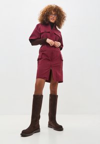 Vestido de algodón burdeos con cierre de cremallera, mangas cortas y dos bolsillos frontales. Combinado con botas de ante marrón hasta la rodilla.