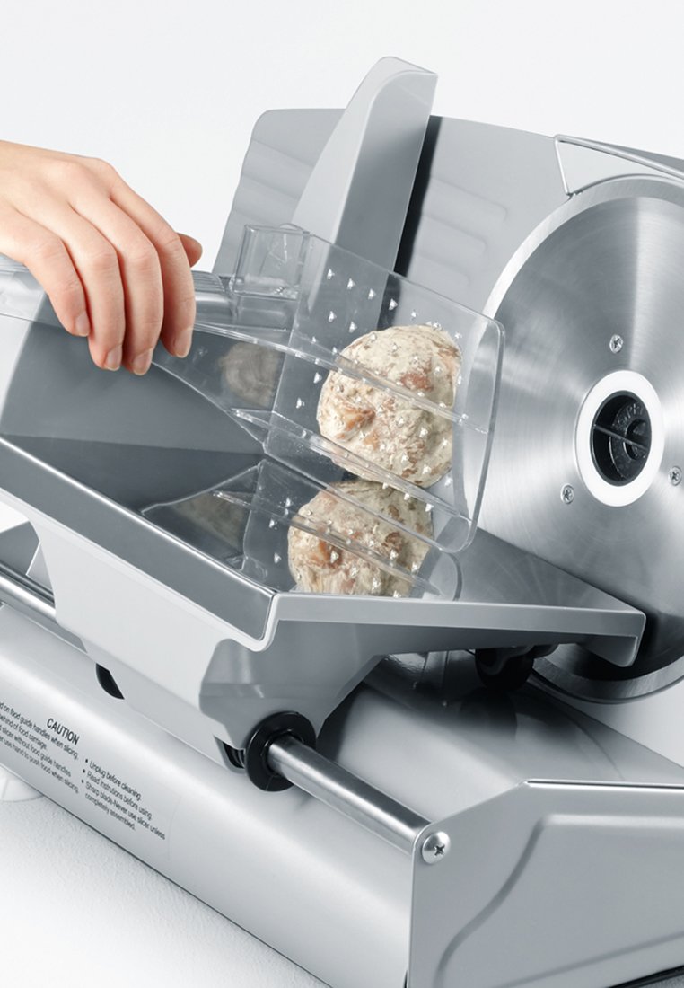 Affettatrice in argento con lama circolare in acciaio inossidabile. Porta cibo in acrilico trasparente in uso, che contiene un pezzo di pasta rotondo.