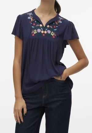 Vrouw draagt een marineblauwe blouse met kleurrijke bloemrijke borduursels op de borst en mouwen met volant, gecombineerd met een donkerblauwe spijkerbroek.