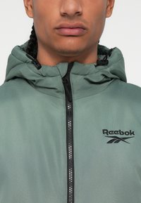Žalios spalvos "Reebok" striukė su užtrauktuku priekyje, gobtuvu ir tekstūruota medžiaga. Yra juodas logotipas ir užtrauktuko akcentai.