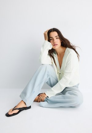 Jonge vrouw met lang bruin haar, dragend een witte blouse, lichtblauwe wijdvallende spijkerbroek en zwarte sandalen, zittend op de vloer tegen een witte achtergrond.