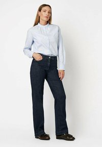 Light blue collared shirt with buttons paired with dark blue straight-leg jeans. Model wearing dark shoes with decorative accents.
