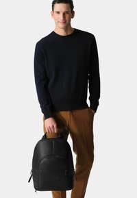 Man wearing a black sweater and brown pants holding a black leather backpack by its top handle against a plain background.