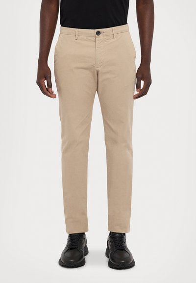 Chinos beiges com corte reto, feitos de algodão, apresentando bolsos laterais e um fecho com botão na cintura. Usados com sapatos pretos.