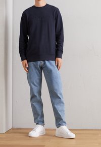 Sudadera azul marino con el texto "Copenhague" en relieve, escote redondo, puños de canalé, combinada con jeans azul claro y zapatillas blancas.