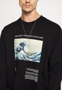 Sudadera negra con el gráfico de "La Gran Ola de Kanagawa" en azul y verde, con texto blanco que detalla la obra debajo. Collar plateado visible.