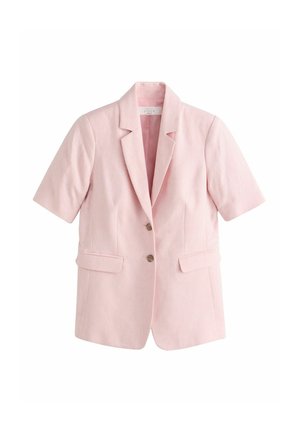 Vrouw draagt lichtroze blazer met korte mouwen en bijpassende broek, staat binnen met de rug naar buiten gericht, met groene bomen zichtbaar door het raam.