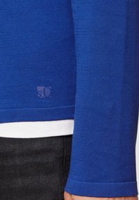 Blauer Strickpulli mit langen Ärmeln, der ein dezentes Logo am Saum aufweist. Glatte Textur, taillierte Passform, über einem weißen Hemd getragen.