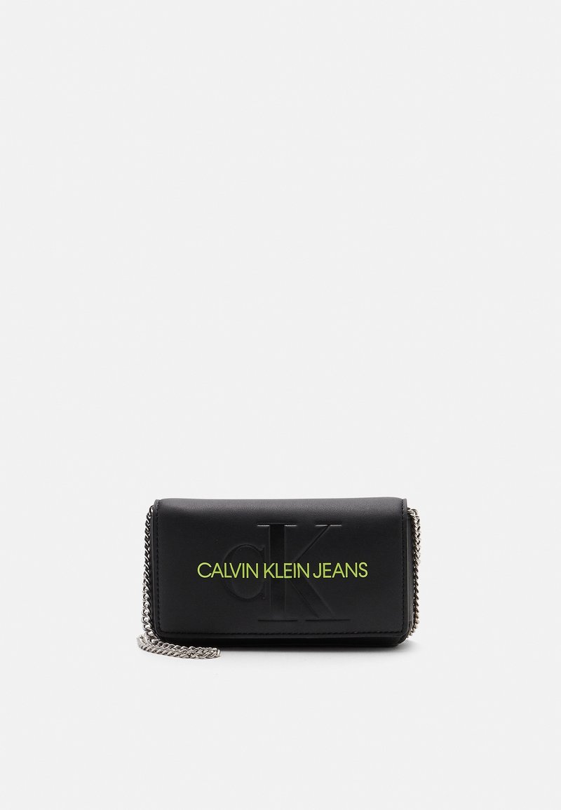 Portafoglio in finta pelle nera con logo embossato, testo neon verde "CALVIN KLEIN JEANS" e tracolla in catena argentata. Forma rettangolare compatta.