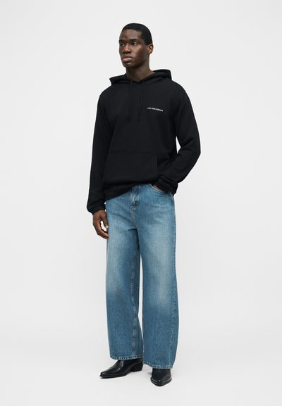 Mann trägt schwarzen Kapuzenpullover mit dem Schriftzug "LES BENJAMINS", weite blaue Jeans und schwarze spitze Stiefel, steht mit einer Hand in der Tasche.