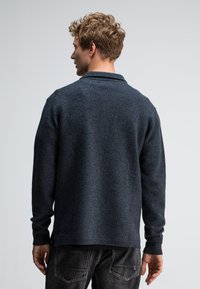 Dunkelgrauer Strickpullover mit Kragen, langen Ärmeln und texturierter Oberfläche. Der Saum ist hinten leicht länger und wird mit dunklen Jeans getragen.