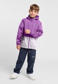 Lila en zilveren lichtgewicht jas met een capuchon en elastische manchetten, gecombineerd met marineblauwe broek. Reflecterende accenten en een logo op de jas.