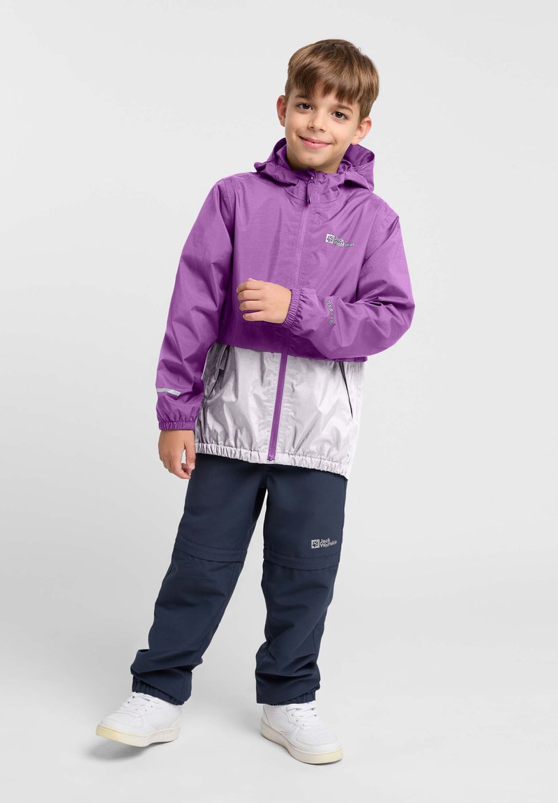 Lila en zilveren lichtgewicht jas met een capuchon en elastische manchetten, gecombineerd met marineblauwe broek. Reflecterende accenten en een logo op de jas.