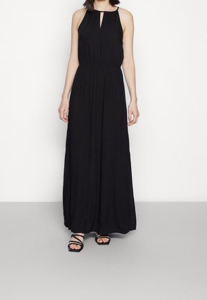 Kobieta w czarnej, bezrękawnej sukni maxi z marszczonym taliowaniem i dekoltem w kształcie łezki, w połączeniu z czarnymi sandałami na ramiączkach.
