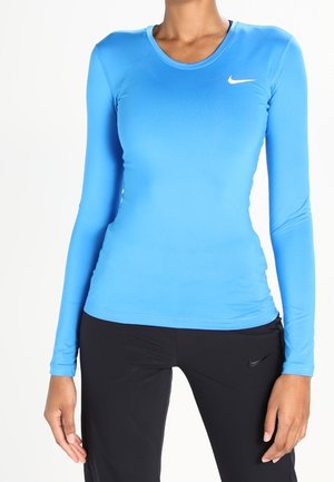 Kobieta ubrana w dopasowaną niebieską sportową bluzkę z długim rękawem Nike i czarne spodnie Nike, stojąca na jednolitym białym tle.