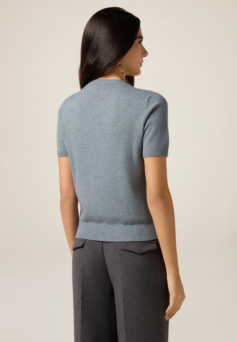 Kortærmet, tætsiddende sweater i blødt blåt uld med ribbet halsudskæring og kant. Kombineret med skræddersyede, mørkegrå bukser med sidelommer.