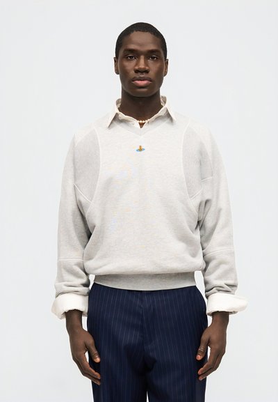 Άνδρας που φοράει ανοιχτό γκρι πουλόβερ πάνω από λευκό πουκάμισο με γιακά και μπλε ναυτικό παντελόνι με ρίγες, στέκεται μπροστά σε απλό φόντο.