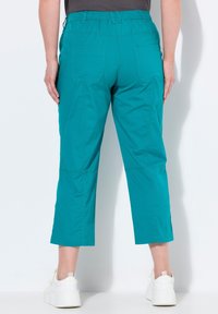 Pantaloni taper in teal con vita elastica, dotati di due tasche posteriori e una texture liscia; abbinati a sneakers bianche.