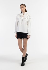 Blusa bianca con scollatura a volant, dettaglio a chiave, maniche lunghe e polsini elasticizzati, abbinata a pantaloncini in denim neri e stivali da combattimento neri.