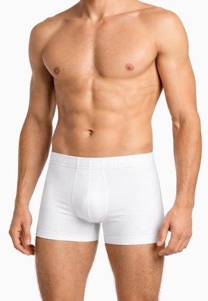 Uomo a petto nudo che indossa boxer bianchi semplici, in piedi con le braccia rilassate ai lati su sfondo bianco.