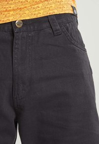 Black denim pants with front button and pocket, paired with orange waistband featuring repeated "Dickies" logo.