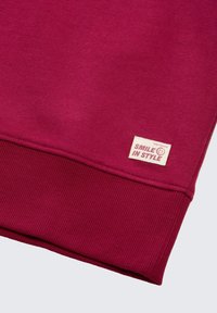 Bordeaux Sweatshirt mit gerippten Bündchen und Saum, versehen mit einem gewebten Etikett mit der Aufschrift "SMILE IN STYLE" und Logo-Detail. Weicher, strukturierter Stoff.