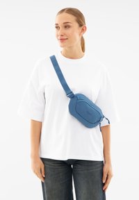 Sininen olkalaukku, joka on valmistettu pehmeästä materiaalista ja jolla on pyöreä muoto, varustettuna vetoketjulla ja säädettävällä hihnalla. Laukkua käytetään valkoisen paidan päällä.