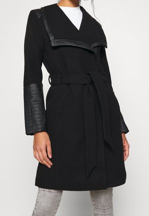 Femme portant un manteau noir ceinturé avec des détails en cuir matelassé sur le col et les manches, associé à un jean gris clair.