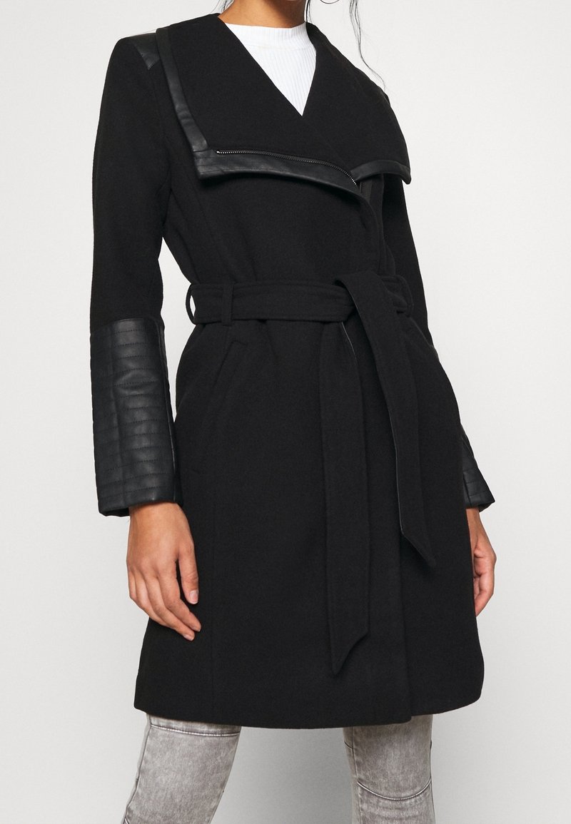 Femme portant un manteau noir ceinturé avec des détails en cuir matelassé sur le col et les manches, associé à un jean gris clair.