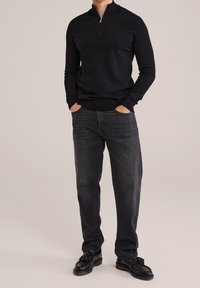 Felpa nera con zip e maniche lunghe, abbinata a jeans grigio scuro e scarpe nere con dettagli a nappine. Texture liscia e aderente.